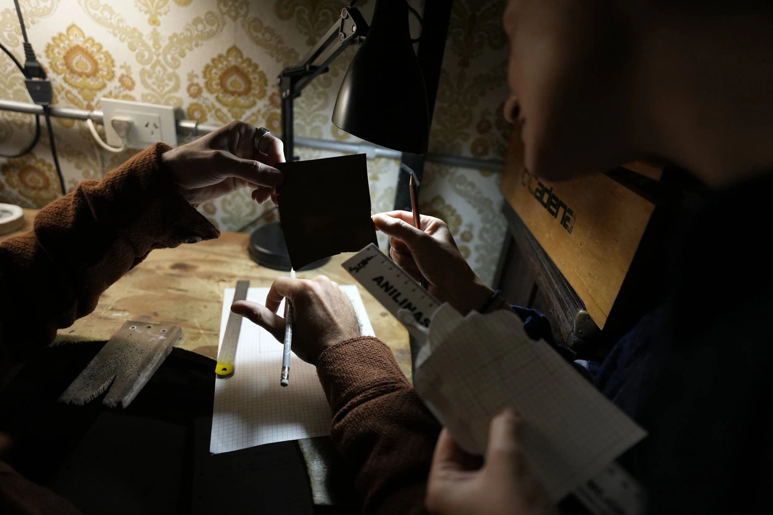 Two people working at a wooden desk, examining photographs, with a notebook, pen, and various office supplies, under a black desk lamp, in a room with floral wallpaper and multiple electrical outlets.