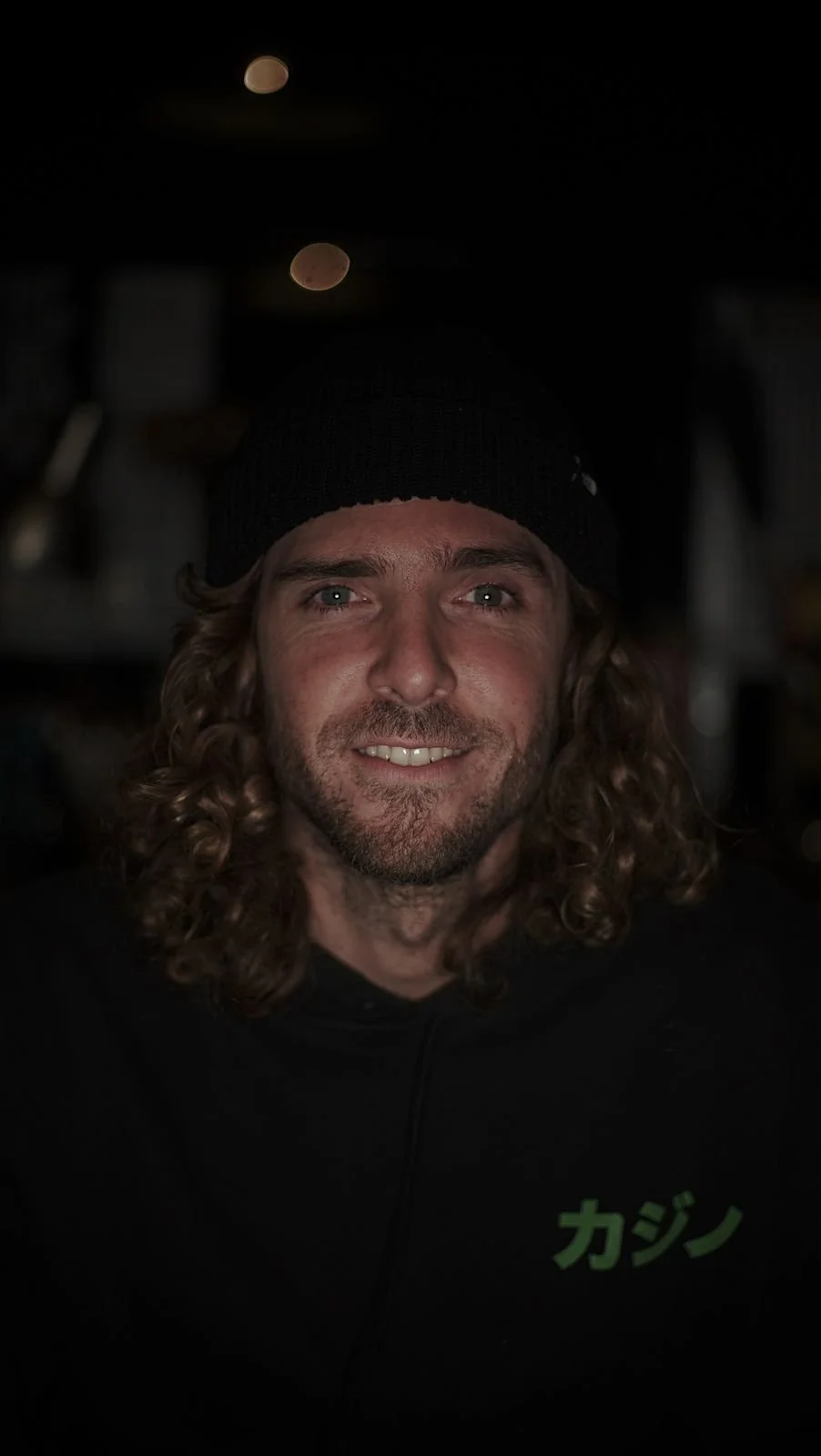 A man with long, curly hair and a beard, wearing a black beanie and black hoodie with Japanese characters, smiling at the camera, with dark background and out-of-focus lights behind him.