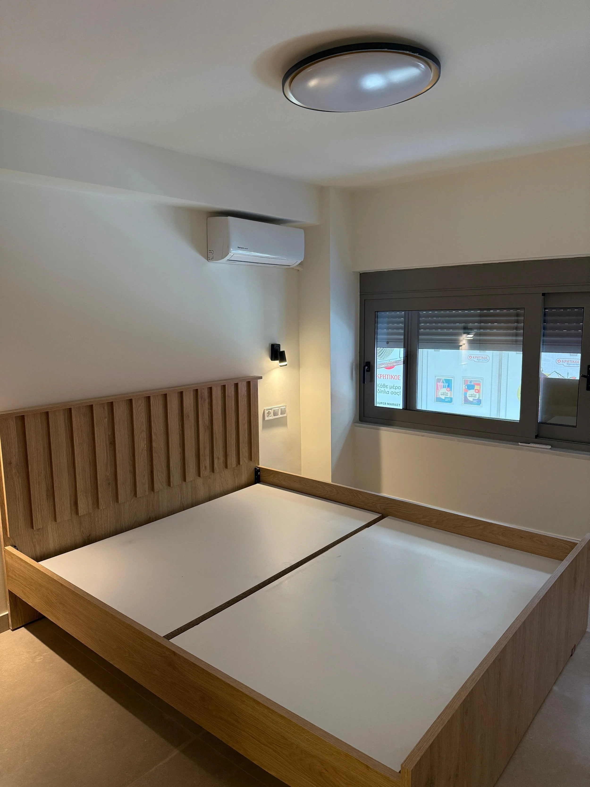 Empty bedroom with a wooden bed frame, a large window, and an air conditioning unit on the wall. Greece, Investment, Golden Visa, Real Estate, Cyprus, Georgia, Lebanon, LPI, Athens.