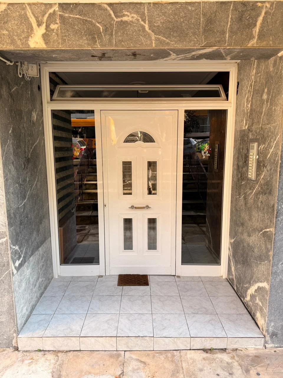 Front entrance with a white door and glass windows, tiled porch, gray marble walls, and a small brown doormat.