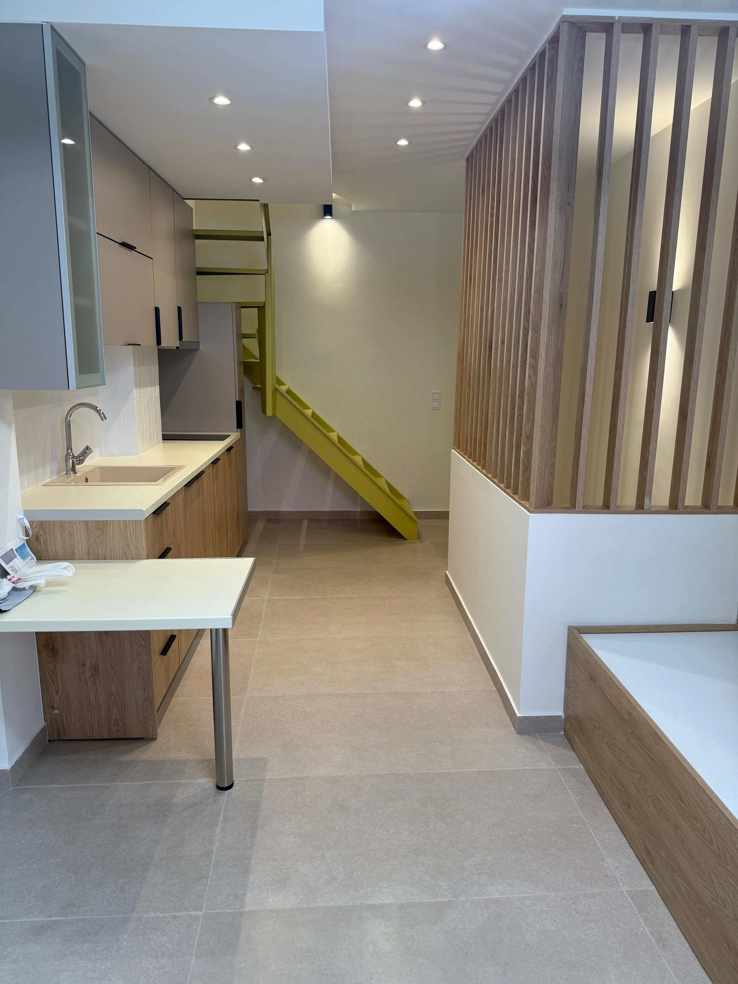 Empty modern kitchen with beige cabinets, a small sink, a yellow staircase in the background, and a wood-paneled partition on the right. Greece, Investment, Golden Visa, Real Estate, Cyprus, Georgia, Lebanon, LPI, Athens.