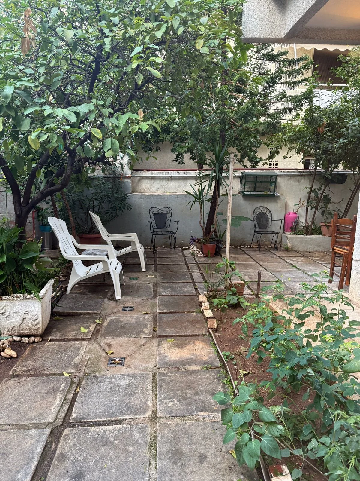 A small, urban backyard patio with large paver stones, surrounded by green shrubs and trees, and furnished with white and black plastic chairs, wooden chairs, and a small garden bed.