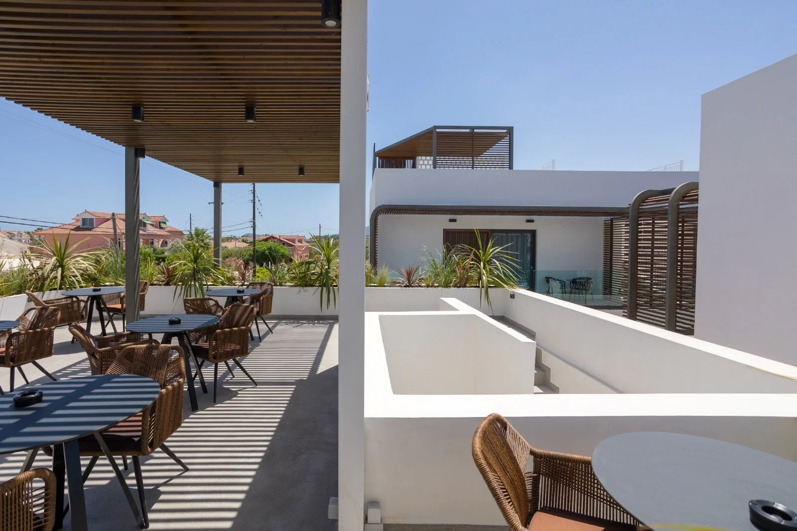 Outdoor patio with wicker chairs and round tables with striped shadows cast by a wooden pergola, plants along the railing, and neighboring modern white buildings under a clear blue sky.