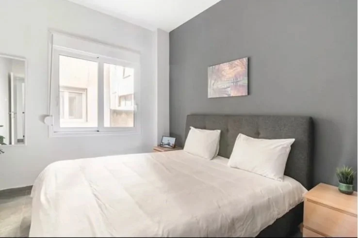 A neatly made bed with white bedding and pillows in a modern bedroom, with a gray accent wall, a small wooden nightstand, a painting above the bed, and a window with blinds.