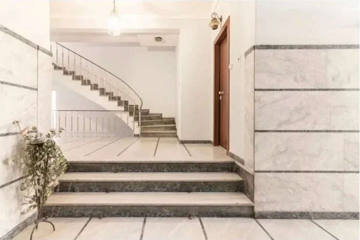 Interior of a building with marble stairs and a curved staircase leading upward, next to a wooden door and a potted plant near the stairs. Greece, Investment, Golden Visa, Real Estate, Cyprus, Georgia, Lebanon, LPI, Athens.