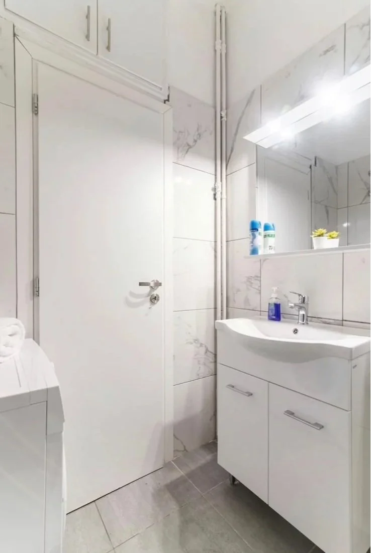 A small modern bathroom with a white sink cabinet, a mirror, and a white door. There are bottles of spray and a plant on the shelf above the sink. The walls are tiled with marble-patterned tiles.