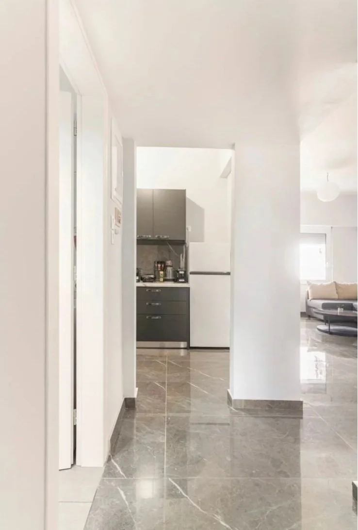 View of a modern kitchen with gray cabinets and a white refrigerator, seen through an open doorway from the hallway, with a living room visible in the background.