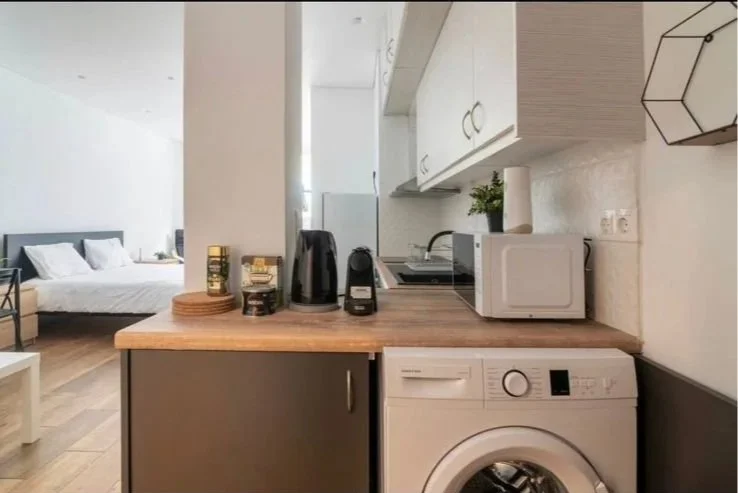Compact kitchen with microwave, toaster, kettle, small plant, and washing machine, adjacent to a bedroom visible in the background.
