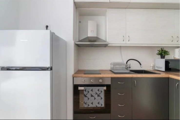 Small kitchen with white cabinets, a refrigerator, a microwave, a dish rack, a stove with an oven, and a sink with a black faucet. There is a small green plant on the countertop.