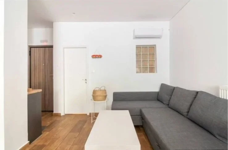 Living room with a gray sectional sofa, a white coffee table, a small side table with a basket, a window with glass blocks, a door, and an air conditioning unit on the wall.