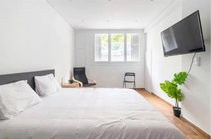 Simple bedroom with a large bed, white bedding, a black chair, a small side table, a window with blinds, a wall-mounted TV, and a green potted plant