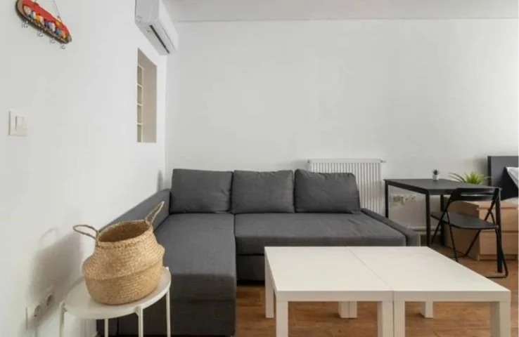 Living room with gray sectional sofa, white coffee table, black dining table with two chairs, wicker basket on a small white side table, air conditioning unit, white walls, and wooden flooring.