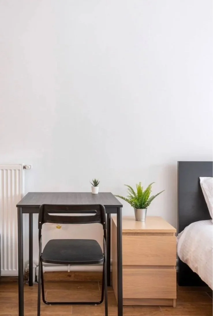 A bedroom with a small black desk, a black chair, a wooden nightstand with a potted fern, a bed with white bedding, and a white wall with a radiator.