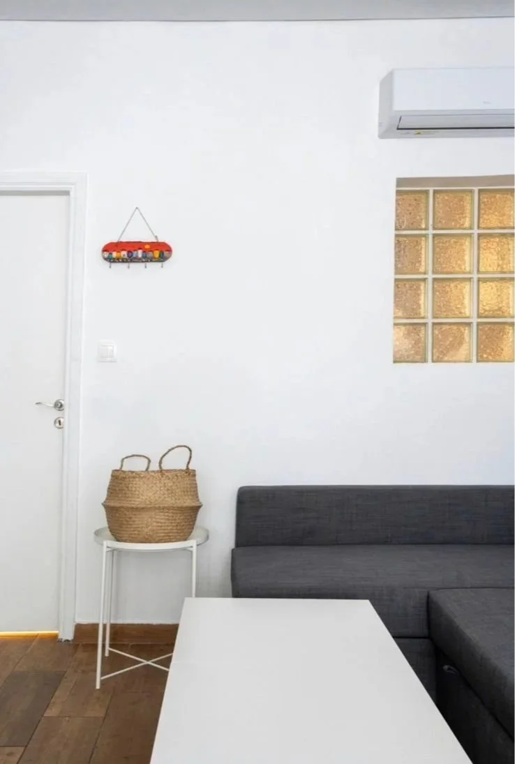 Living room corner with a grey sofa, white side table holding a woven basket, white wall, glass block window, air conditioner, and a decorative wall hanging with a red background