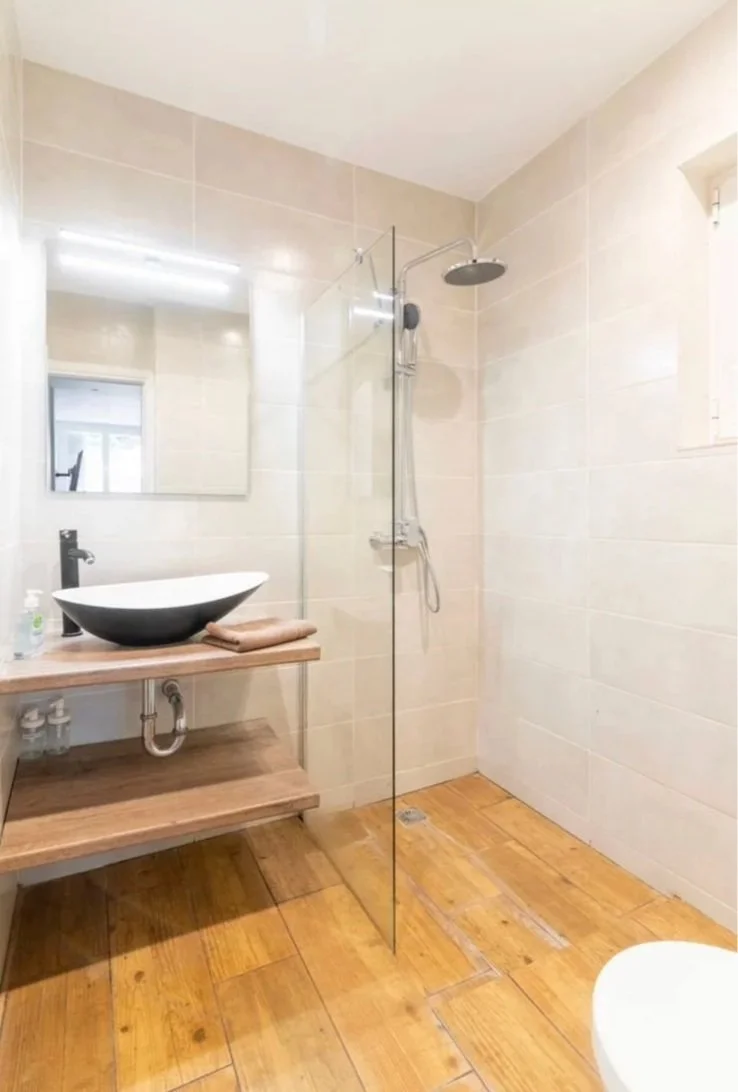 Modern bathroom with a wooden floor, floating wooden vanity with a vessel sink, mirror, and a glass shower enclosure, beige tiled walls, and a small window.