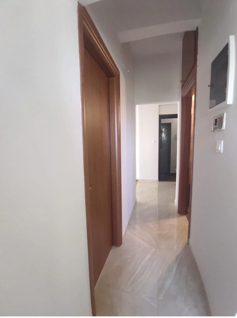 Hallway with wooden doorframes and glossy beige tile floors, leading to a room with a black door at the end.