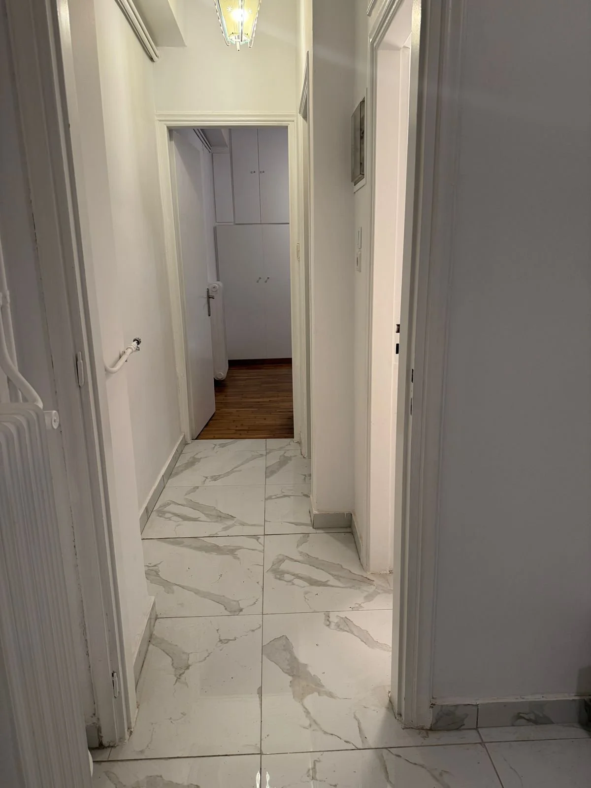 Hallway with white marble floor tiles and white walls, leading to a room with wood flooring and built-in cabinets.