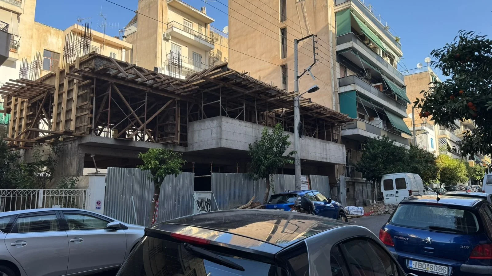 Construction site of a building in an urban area, surrounded by parked cars. The building's framework and scaffolding are visible, with neighboring apartment buildings and a clear blue sky in the background.