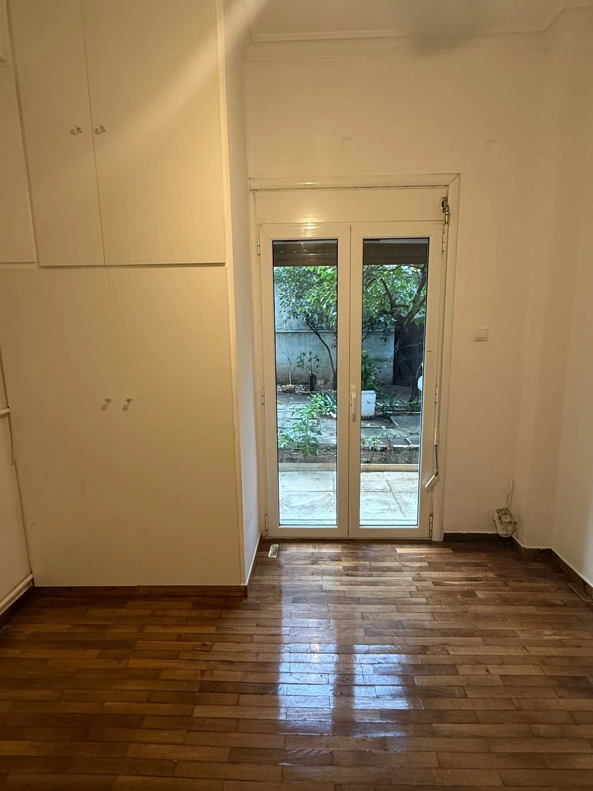 A room featuring an open glass door leading to a garden. The room has wooden flooring and white walls, with a built-in white cabinet on the left side.