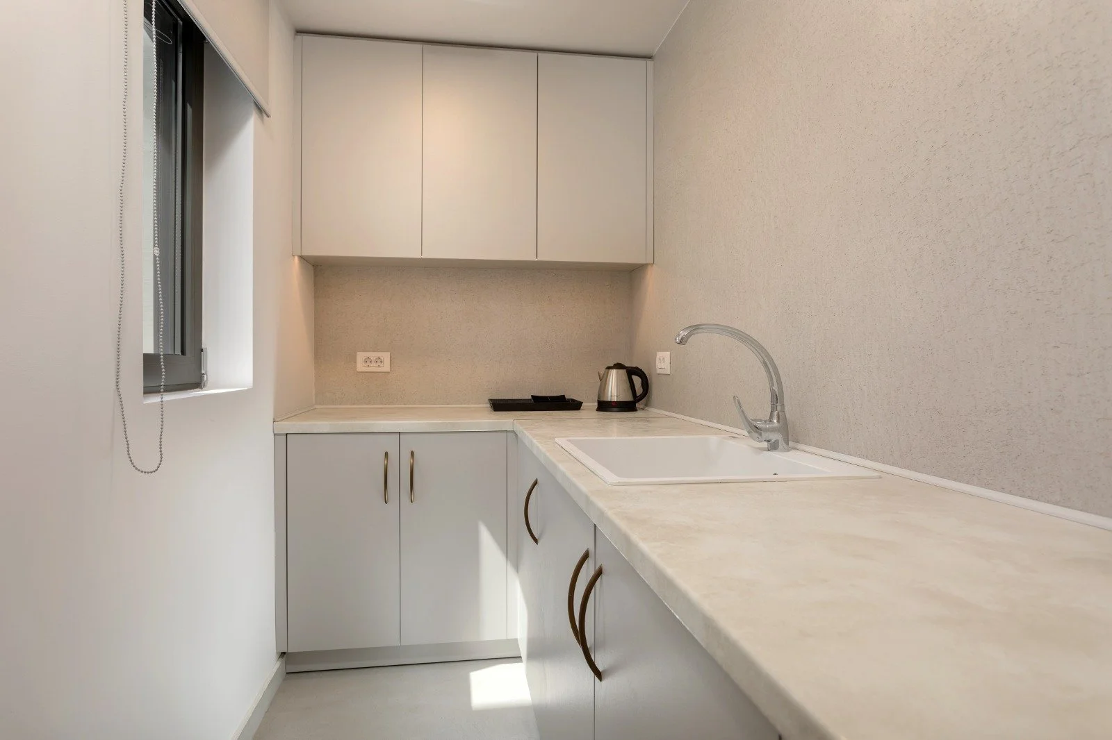 A small, minimalist kitchen with white cabinets and a beige countertop. There is a window on the left side with a chain for blinds. On the countertop, there is a kettle and a black tray. The wall behind the counter has two power outlets, and beige te