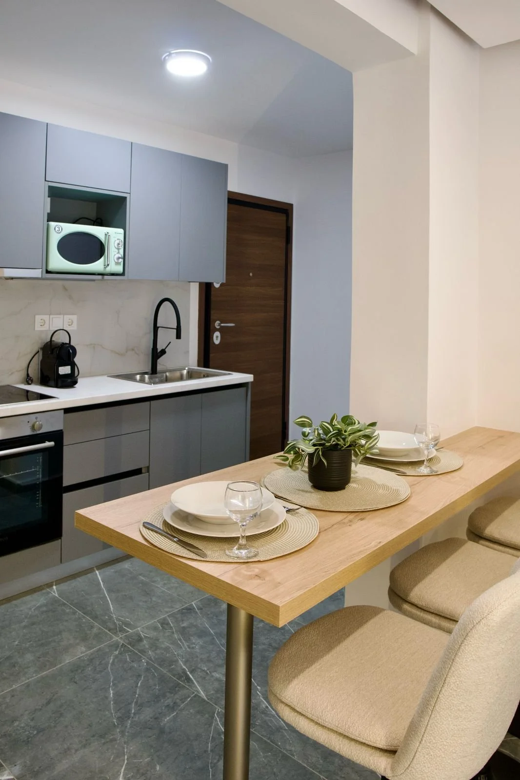 Modern kitchen with grey cabinets, a microwave, and a countertop with a sink, adjacent to a dining table set with placemats, plates, wine glasses, and a potted plant. Greece, Investment, Golden Visa, Real Estate, Cyprus, Georgia, Lebanon, LPI, Athens