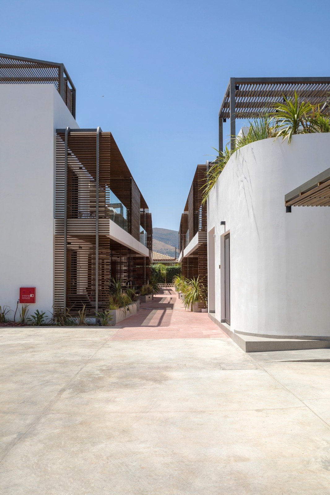 A modern apartment complex with white curved walls, wooden slatted balconies, and potted plants along a walkway, under a clear blue sky.