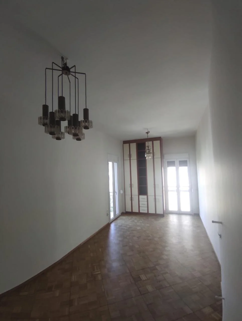 Empty room with wooden flooring, two chandeliers, and glass doors leading to a balcony. There is a built-in cabinet and a window in the background.