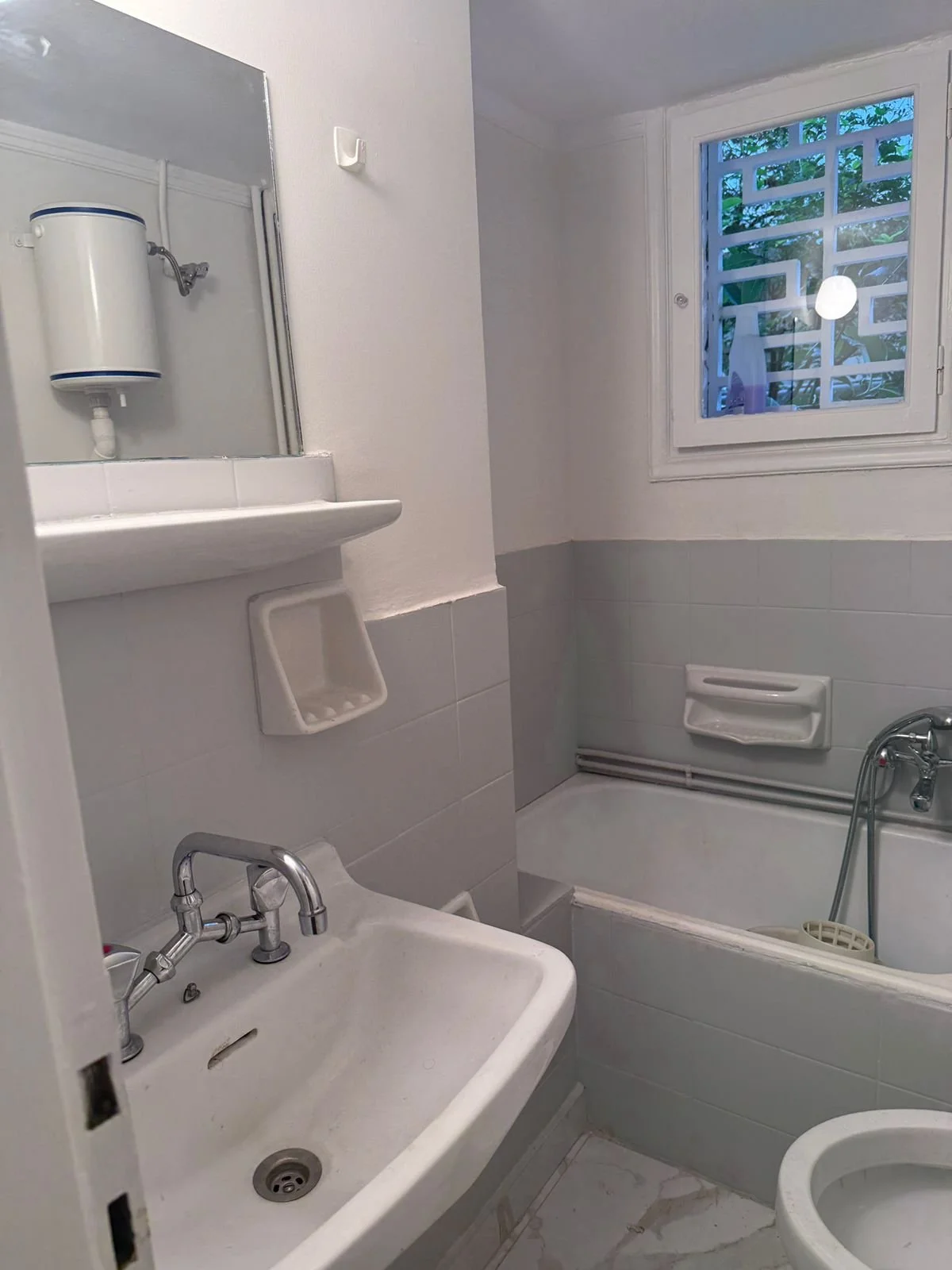 Small bathroom with a sink, mirror, bathtub, window showing greenery outside, and a toilet