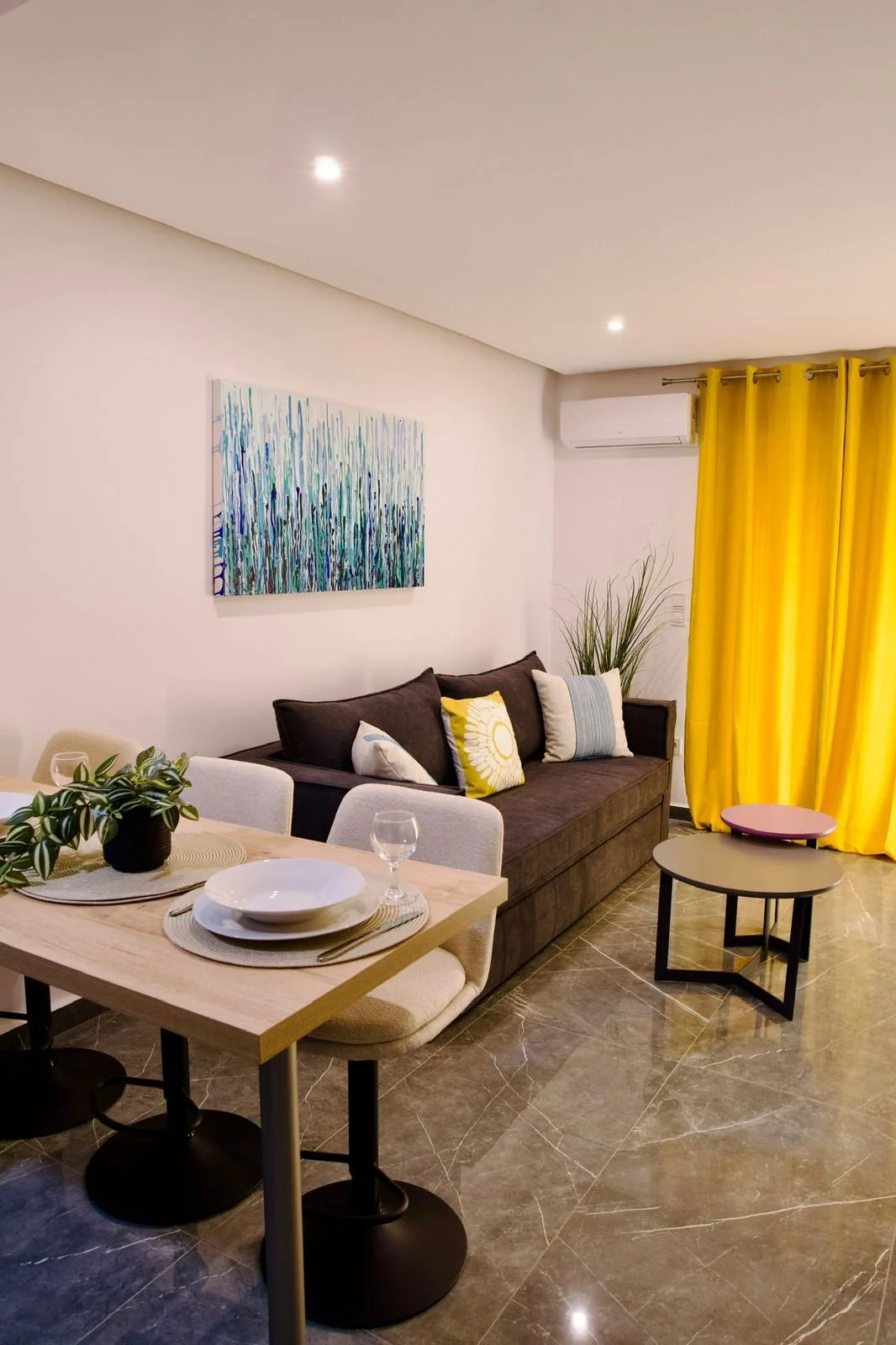 Living room with brown sofa, yellow curtains, abstract wall art, dining table with plates, glasses, and plant, marble floor. Greece, Investment, Golden Visa, Real Estate, Cyprus, Georgia, Lebanon, LPI, Athens.