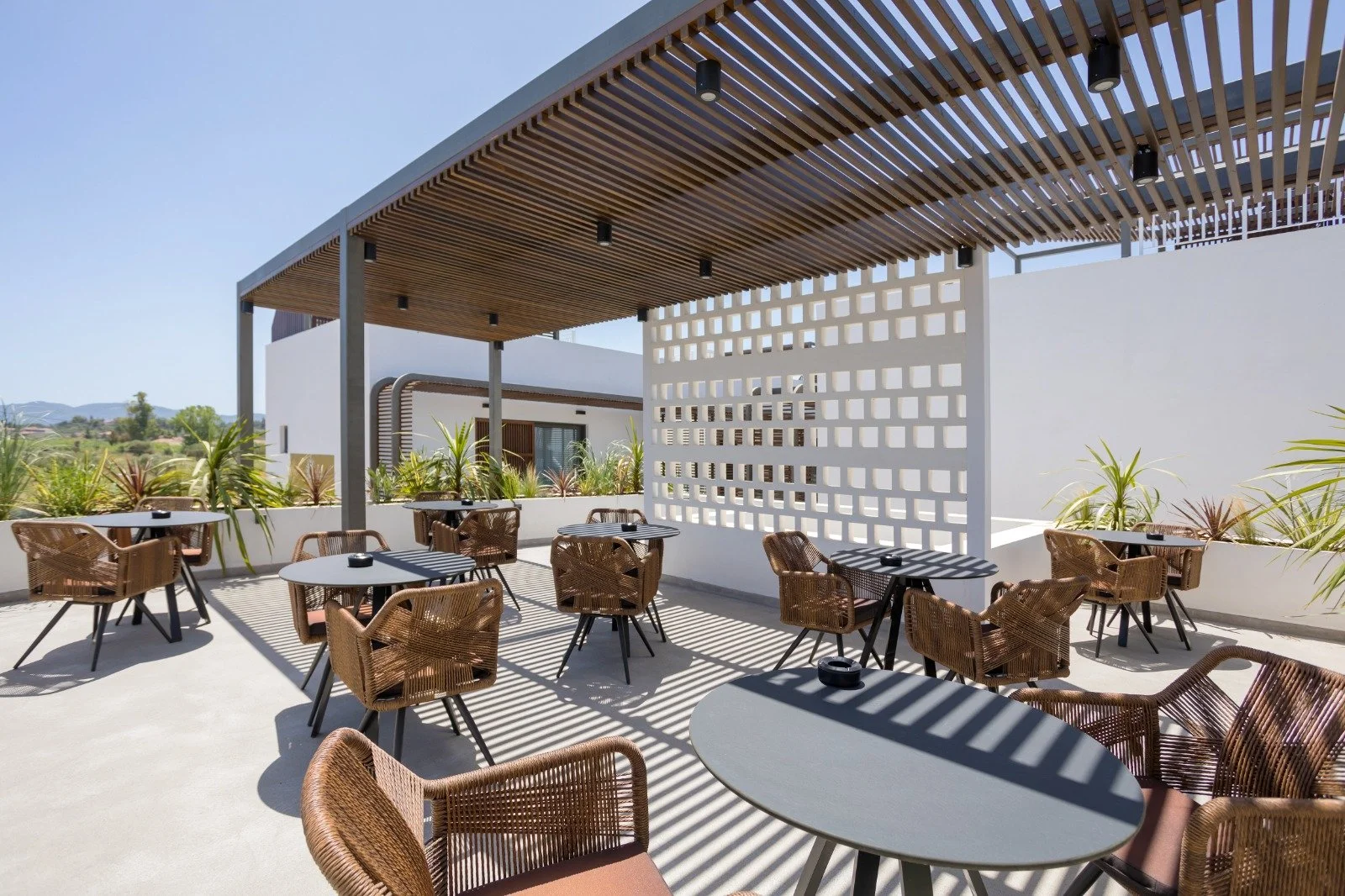 Outdoor patio with wicker chairs and round tables, shaded by a wooden pergola with slats, white decorative wall with square cutouts, potted plants along the perimeter, and a clear blue sky.