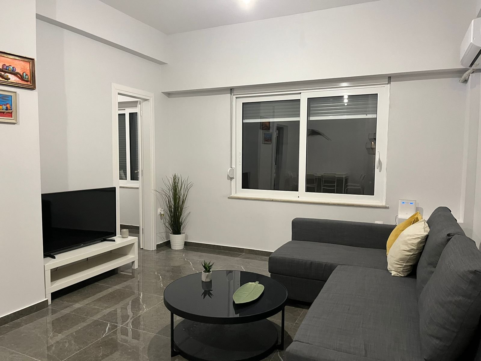 Modern living room with a dark gray sectional sofa, a black round coffee table, a flat-screen TV on a white stand, a potted plant, and decorative pillows. Large window with white frame and a view of a dining area.