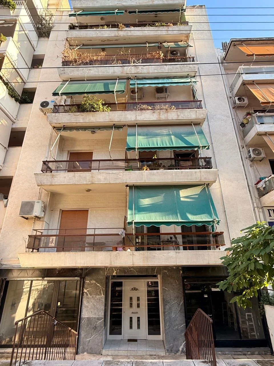 A multi-story residential building with balconies and green awnings, beige exterior walls, some air conditioning units, and a ground floor entrance with glass doors.