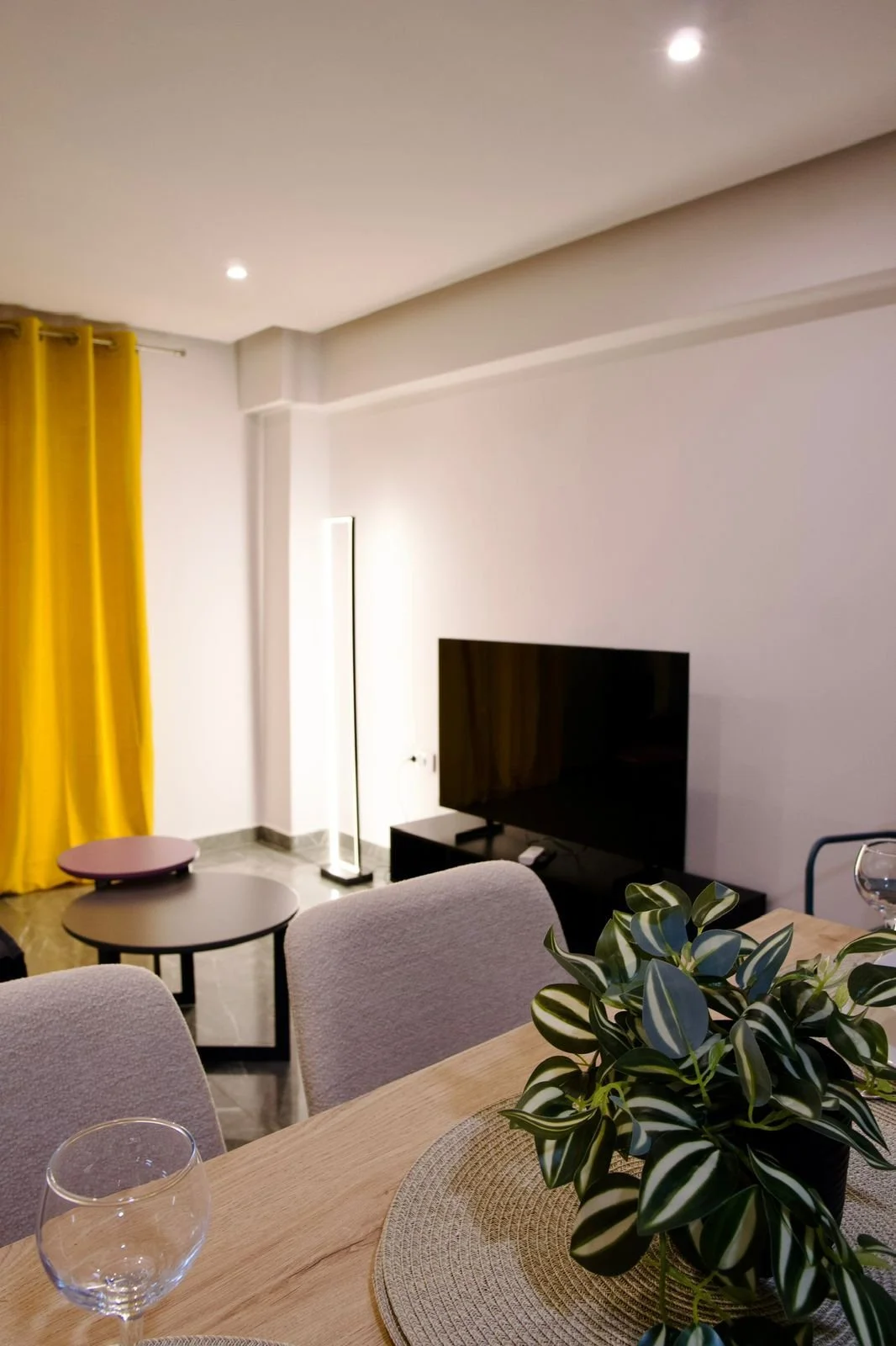 Living room with a mounted TV, floor lamp, yellow curtains, and table with a plant and glasses. Greece, Investment, Golden Visa, Real Estate, Cyprus, Georgia, Lebanon, LPI, Athens.
