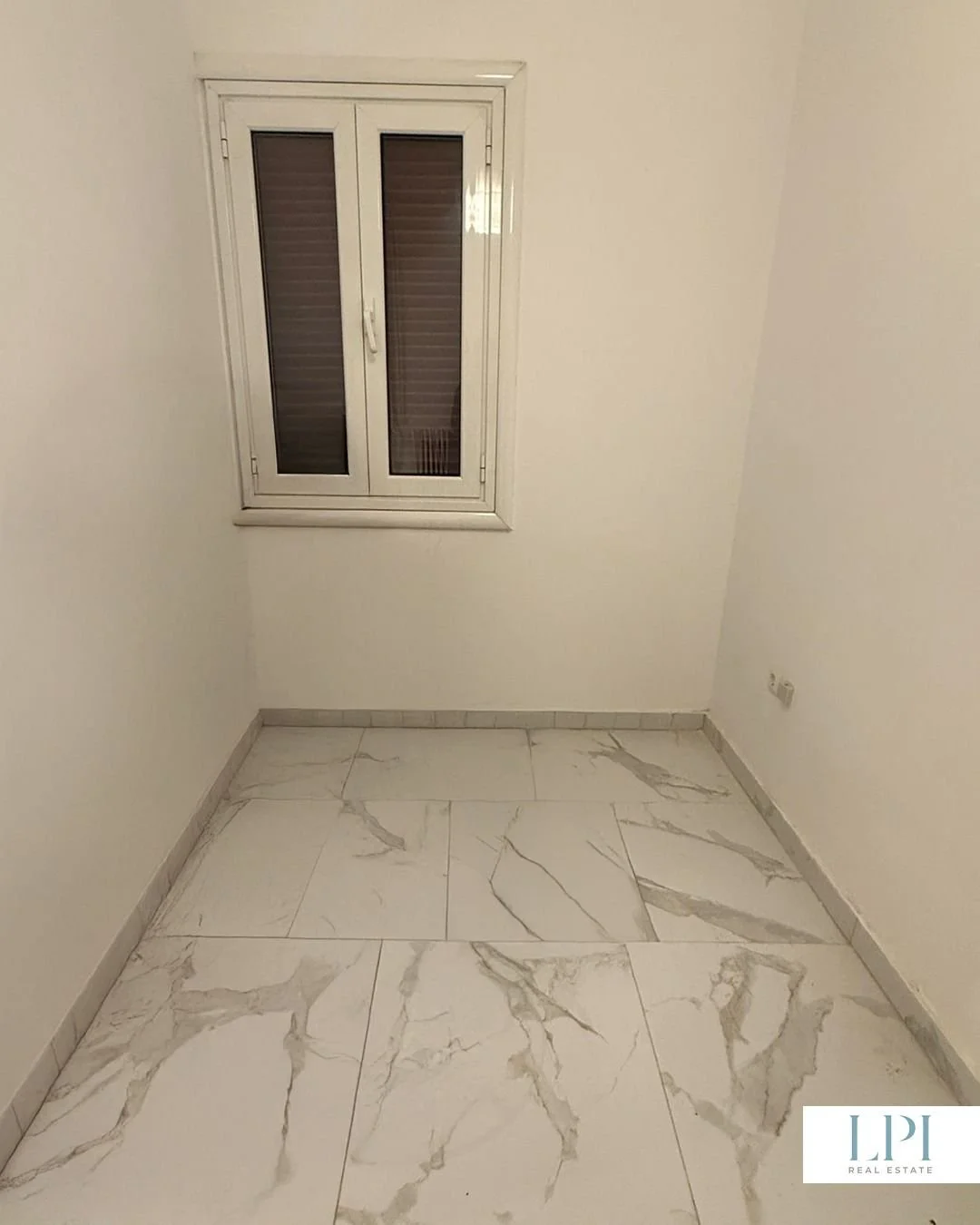 Empty room with white walls, marble tile floor, and a small window with closed blinds.