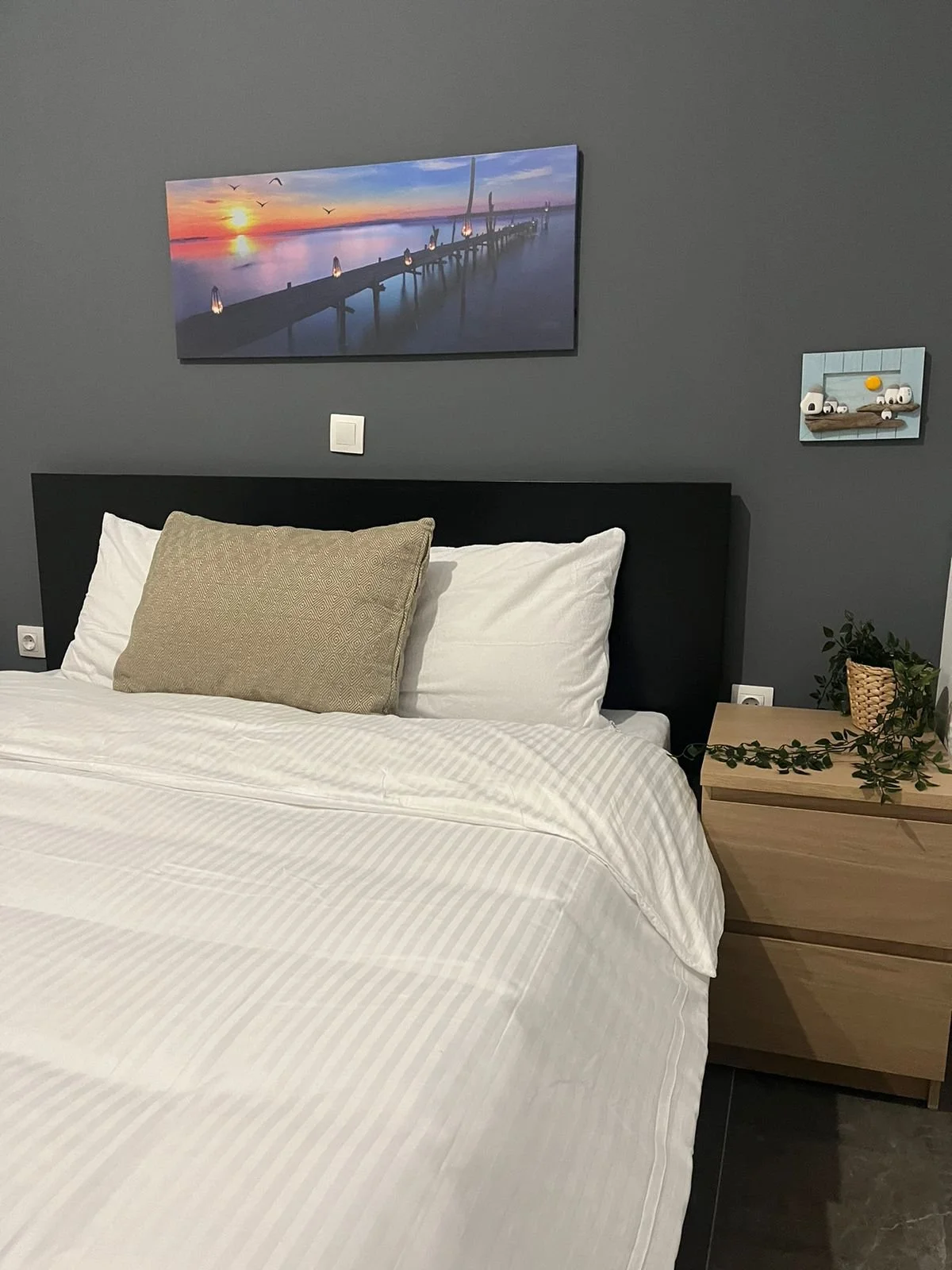 A bedroom with a black headboard, white bedding, and a beige pillow. There is a wooden nightstand with green plants. Two paintings are on the gray wall, one of a sunset over a pier and water, and a smaller one of toy cars.