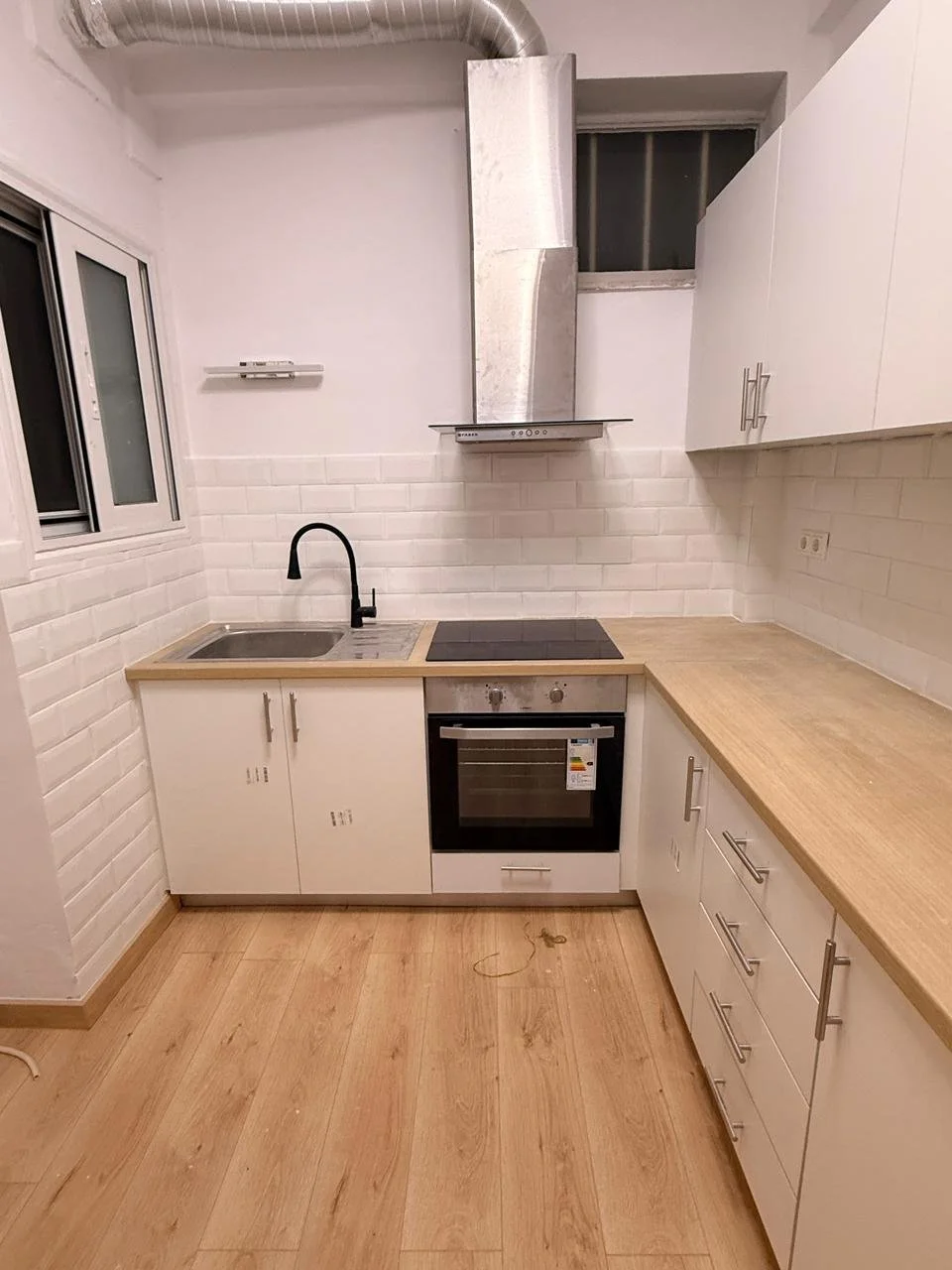 A small modern kitchen with white cabinets, a wooden countertop, a black faucet, a stainless steel oven, and a black electric cooktop. There is a window on the left wall, white tile backsplash, and a small shelf above the sink. The floor is wooden, a