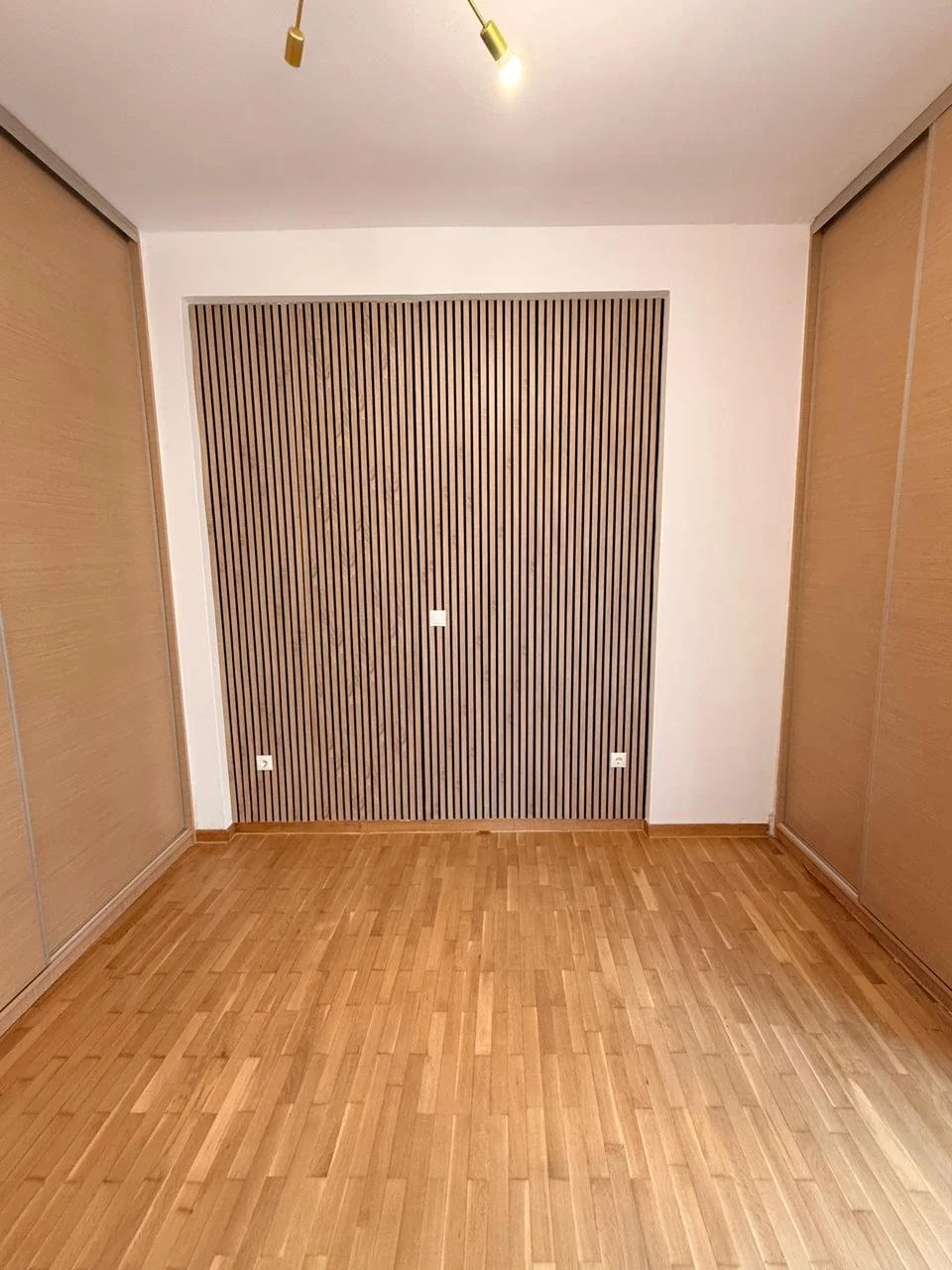 Empty room with wooden flooring, two built-in closets with sliding doors, and a wall with vertical wooden slats. Overhead lighting fixture with two spotlights.