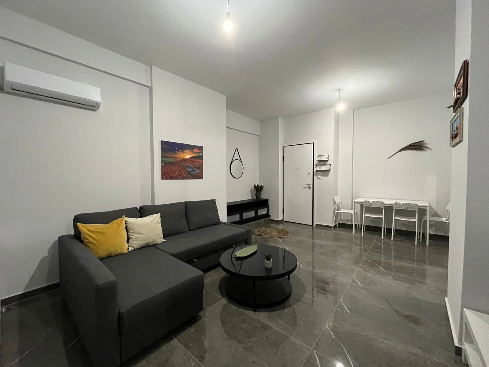 Living room with gray sofa, yellow and beige pillows, black coffee table, white dining table with chairs, wall art, and plant decorations.