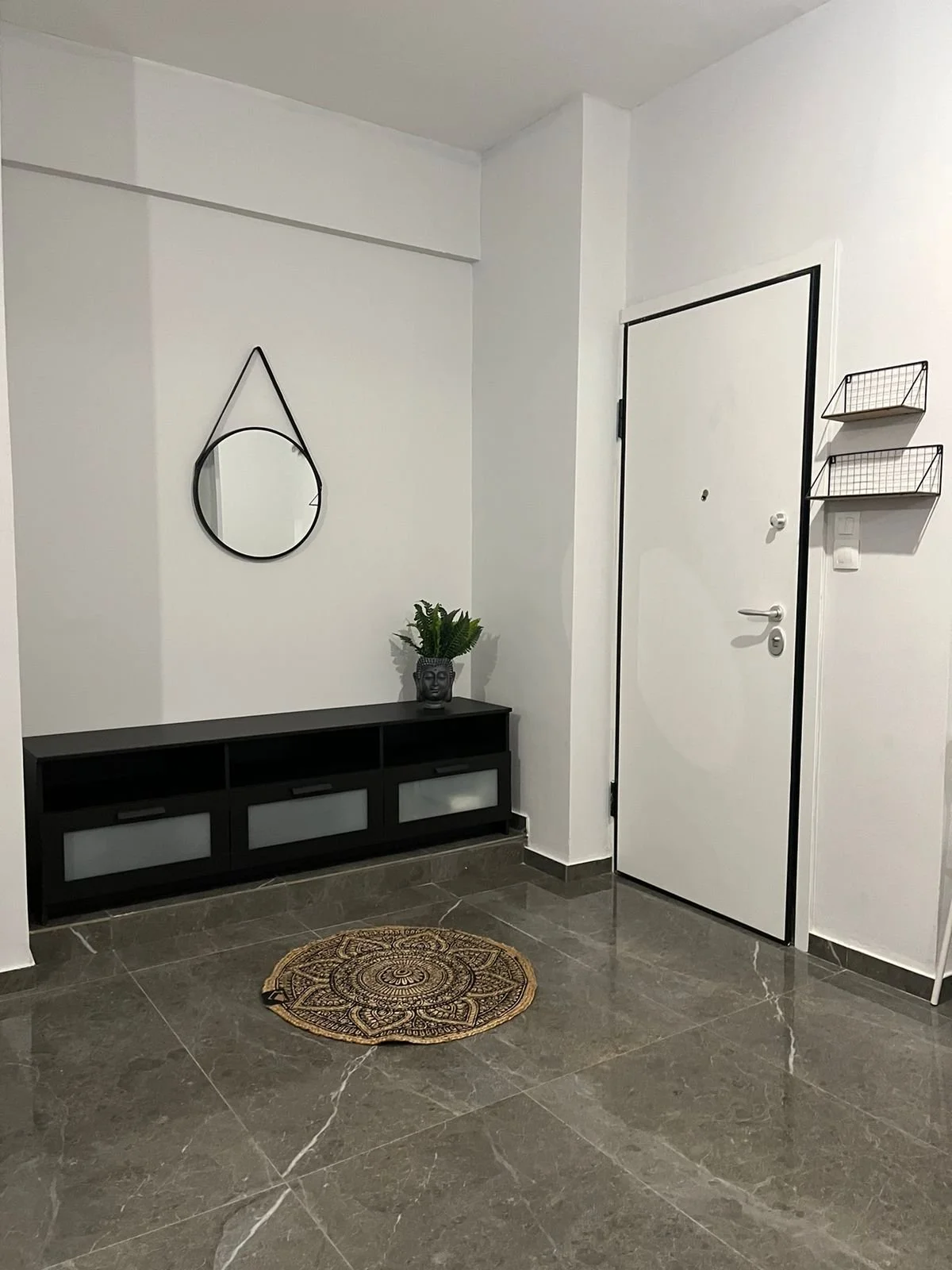 Minimalist entryway with a white door framed in black, a black console table with a plant and decorative object, a round wall mirror, wire wall shelves, and a patterned rug on gray marble floor.
