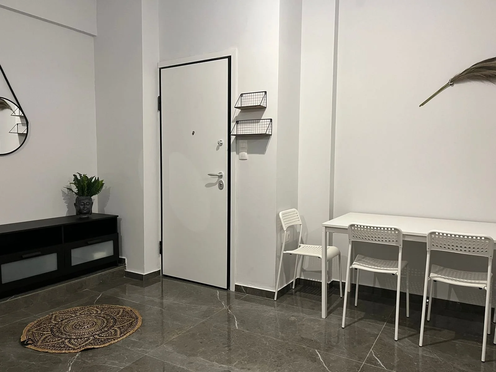 Modern minimalist interior with white walls, white table, white chairs, black console table, a potted plant, and a small decorative rug on a dark tiled floor.