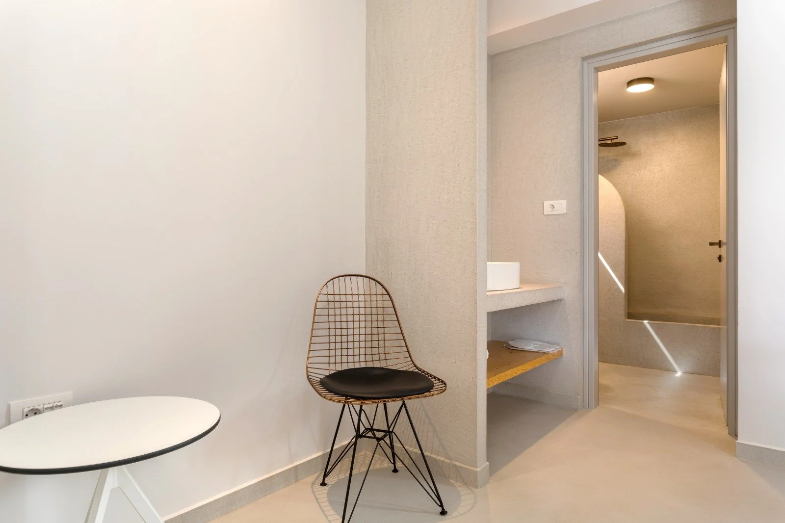 Interior view of a modern, minimalist room with a black wire chair with a cushion, a small leaning white table, and a doorway leading to a shower area with a rain shower head and a circular light on the ceiling.