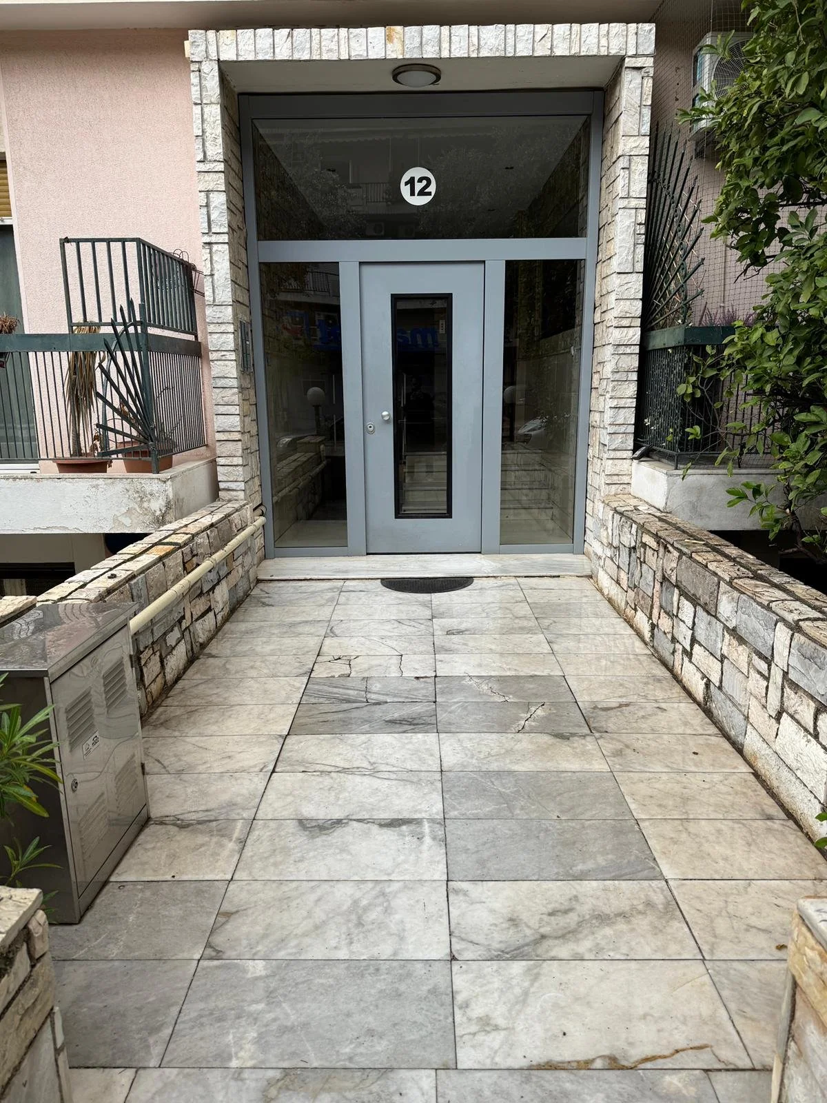 Apartment building entrance with glass door numbered 12, marble tile walkway, brick walls, and greenery. Greece, Investment, Golden Visa, Real Estate, Cyprus, Georgia, Lebanon, LPI, Athens.