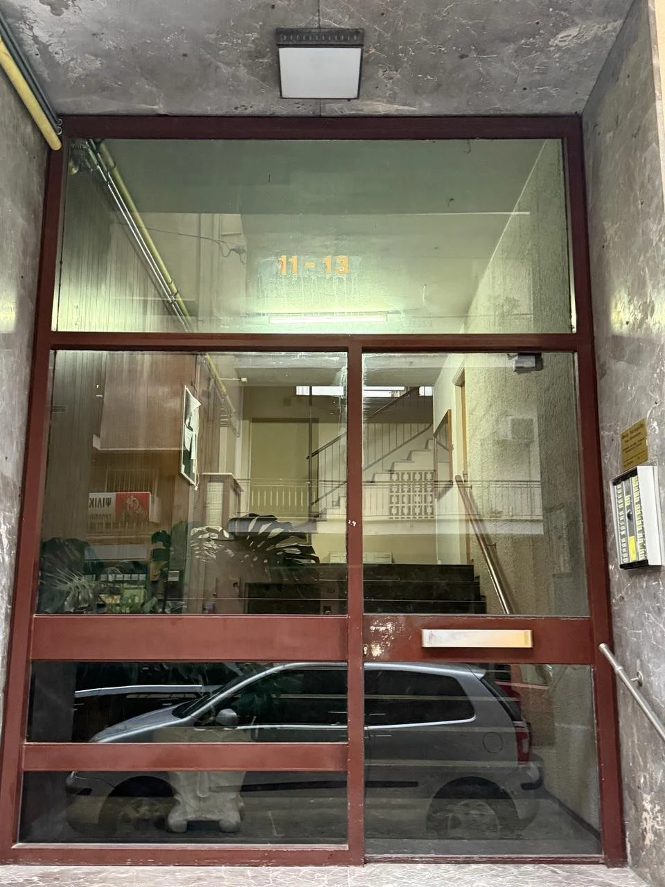 View through glass doors showing an indoor staircase, plants, and a vehicle parked outside. Text '11-13' is visible on the upper glass panel. Greece, Investment, Golden Visa, Real Estate, Cyprus, Georgia, Lebanon, LPI, Athens.