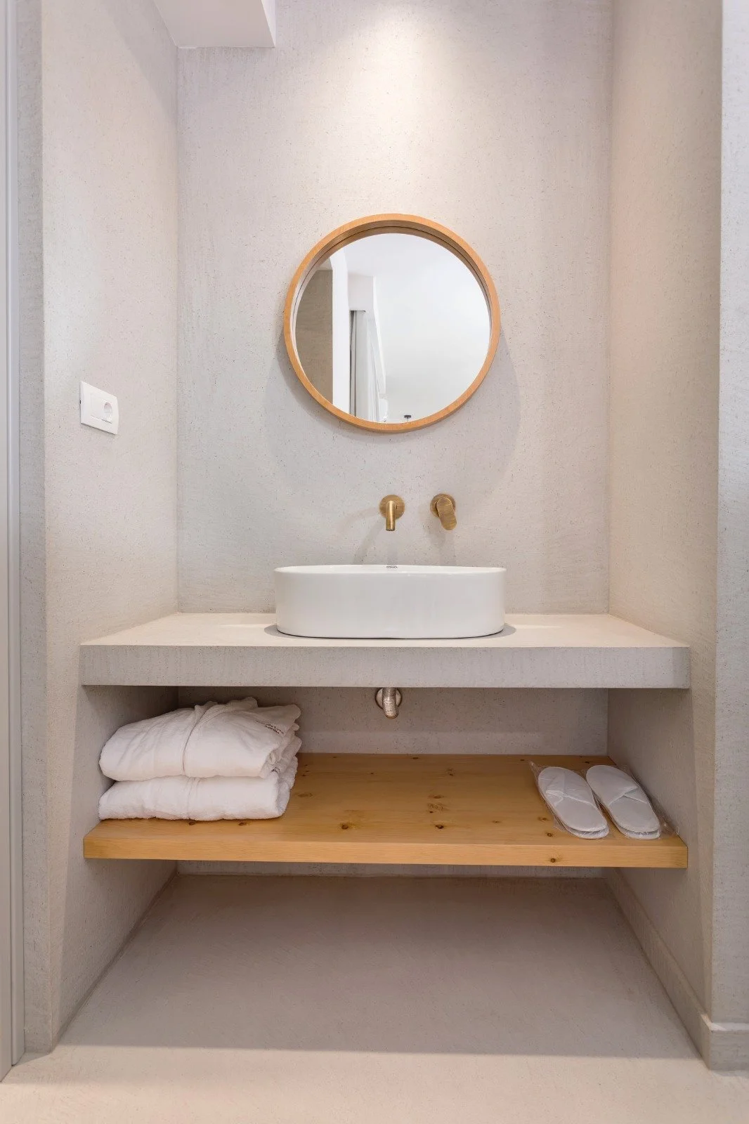 Minimalist bathroom with a round mirror, white sink, gold fixtures, and wooden shelf holding folded towels and slippers.