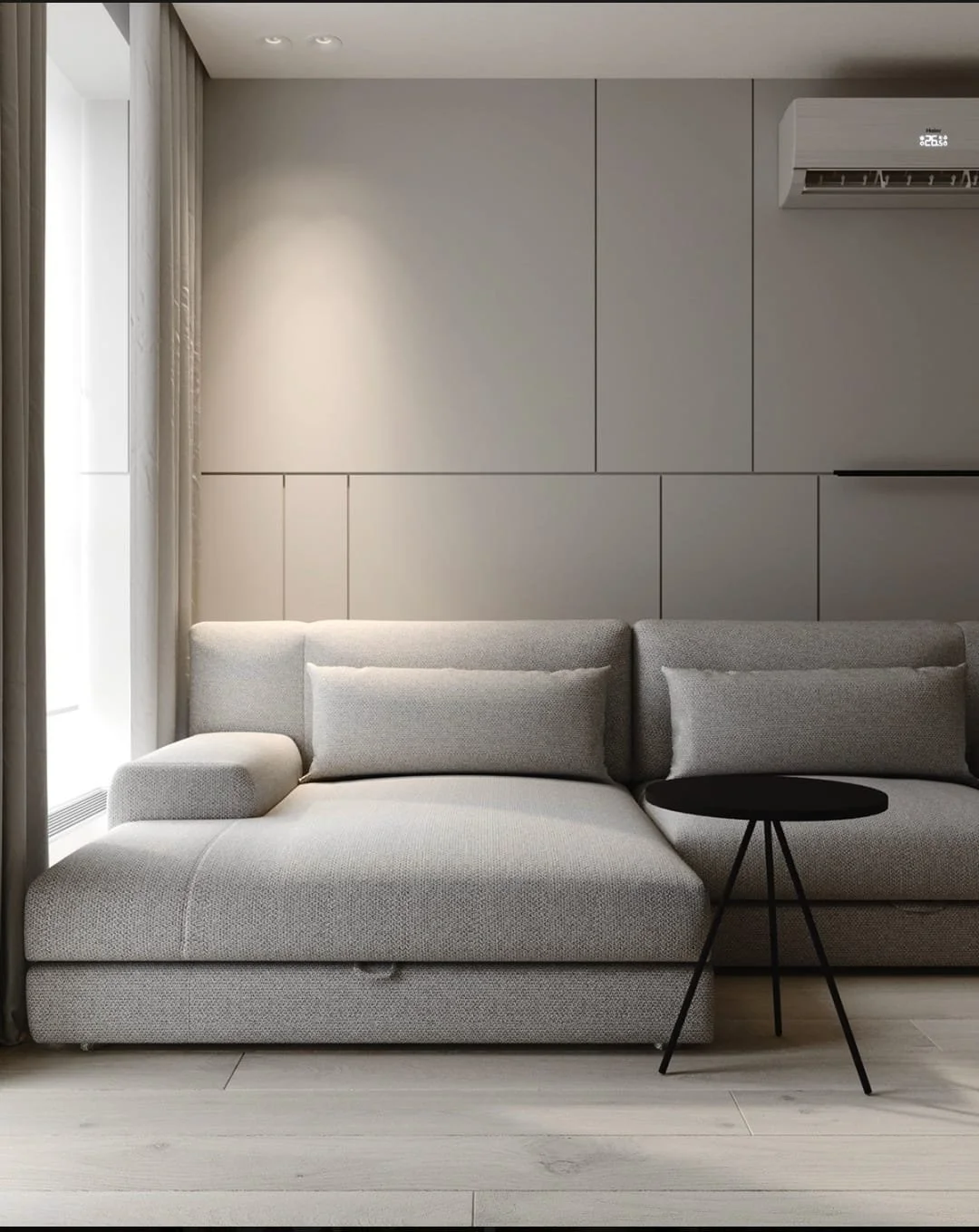 Modern living room with a gray sectional sofa having matching pillows, a round black side table, beige curtains, light-colored flooring, and a wall-mounted air conditioner.