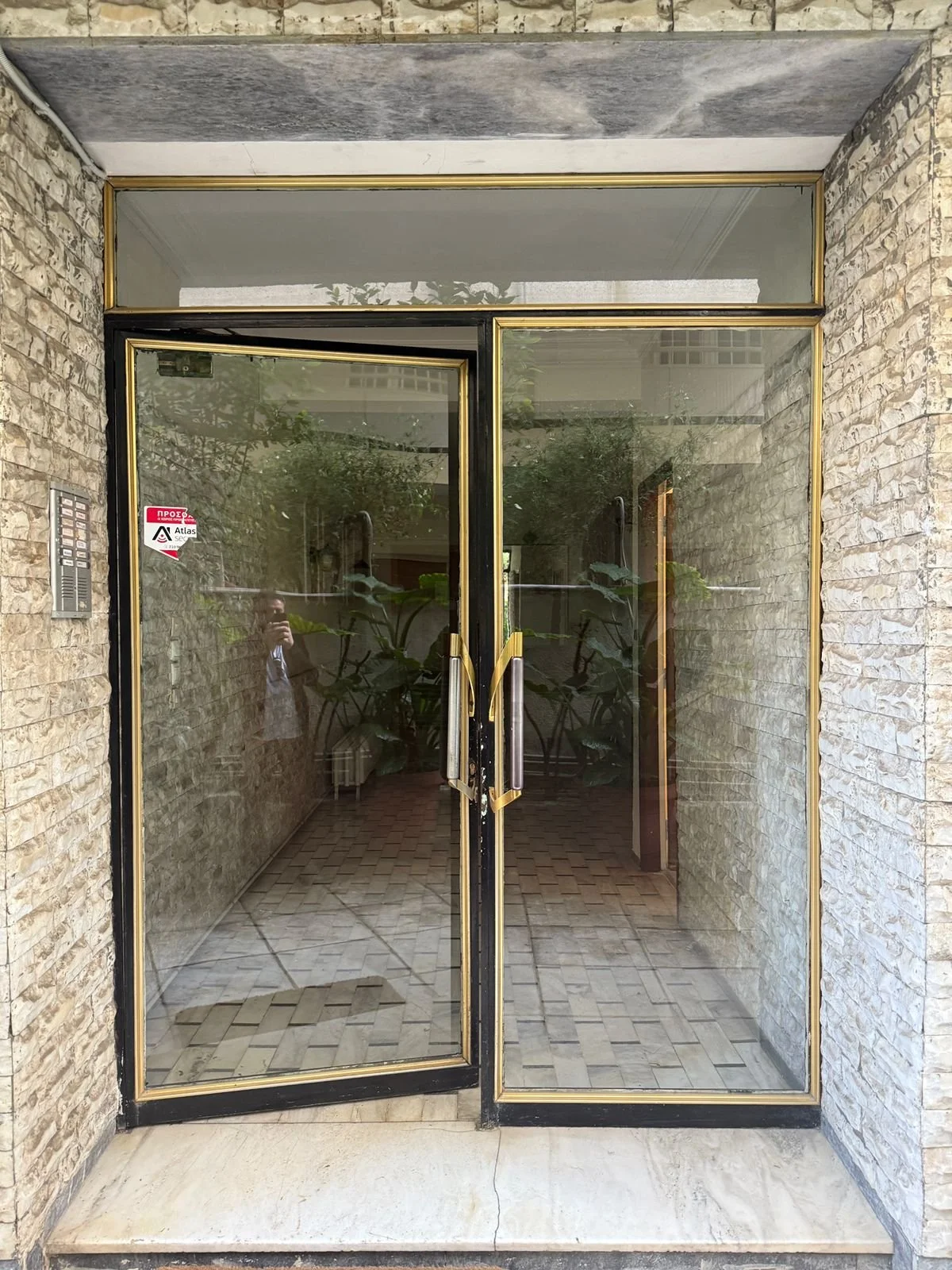Glass door with gold trim and handle, surrounded by stone walls, with a brick-paved interior and plants visible through the glass.