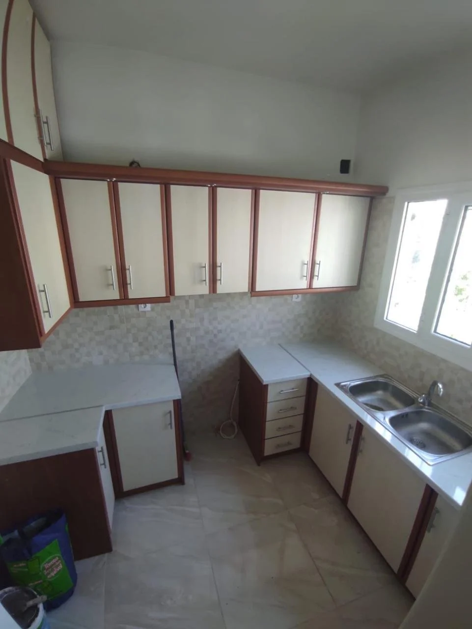Empty kitchen with beige and brown cabinets, a double sink, and a window over the sink, with some cleaning supplies on the floor.