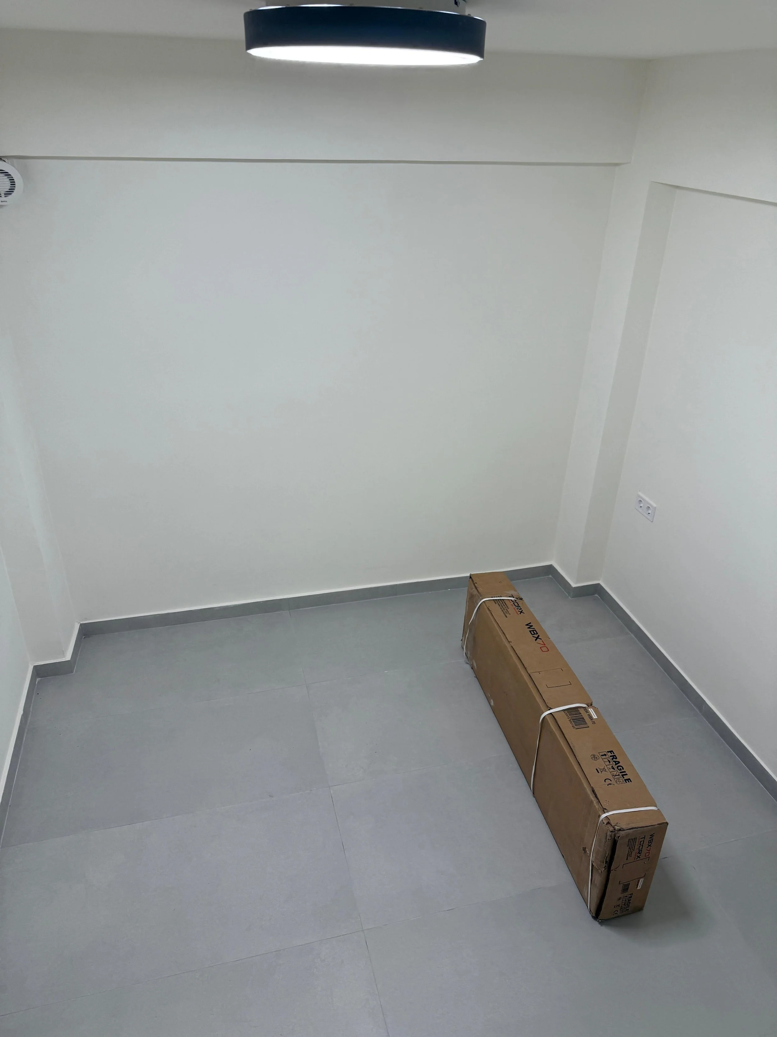 Empty white room with gray tiled floor, a cardboard box on the floor, and a ceiling light. Greece, Investment, Golden Visa, Real Estate, Cyprus, Georgia, Lebanon, LPI, Athens.