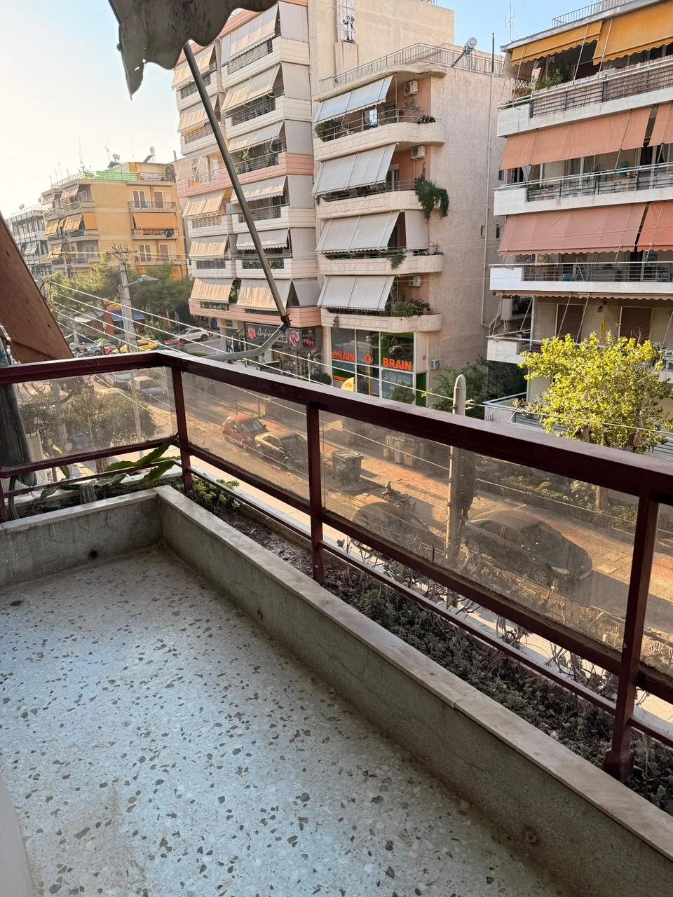 View from a balcony showing a city street with parked cars and multi-story residential buildings with balconies and awnings.