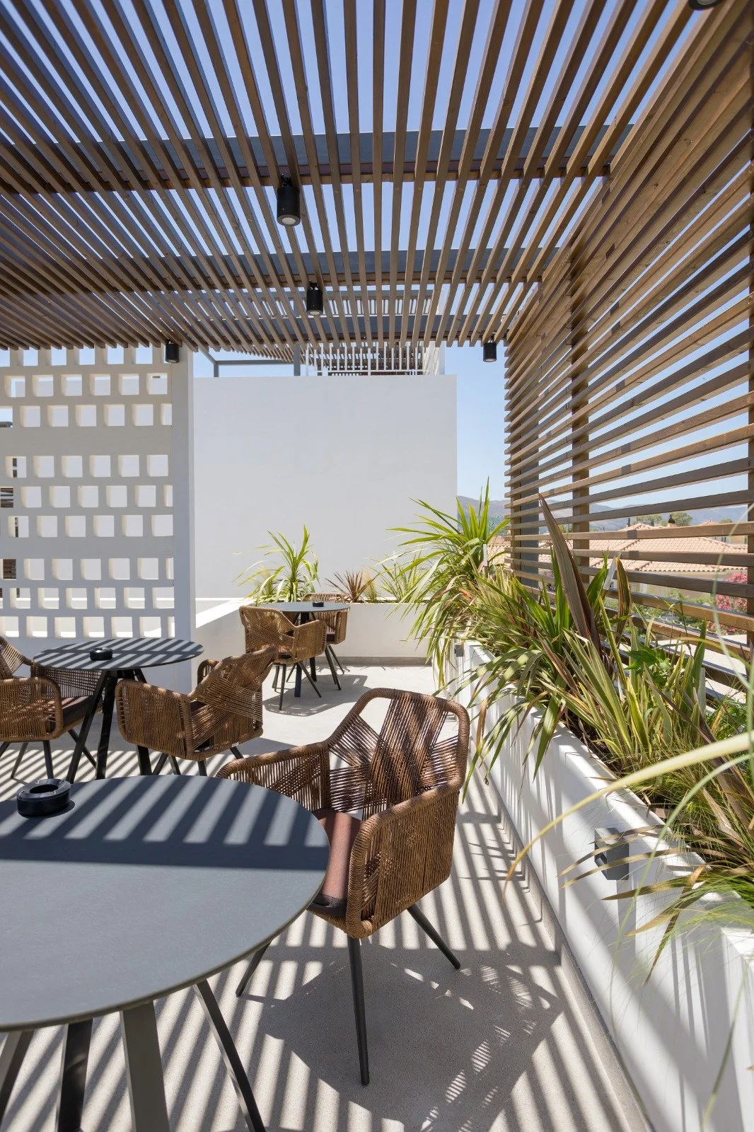 Outdoor patio area with round gray tables, brown wicker chairs, and potted plants. Wooden slat roof and walls create striped shadows on the floor.